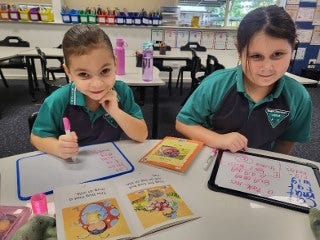 2 students working at desk with mini whiteboards and smiling at the camera